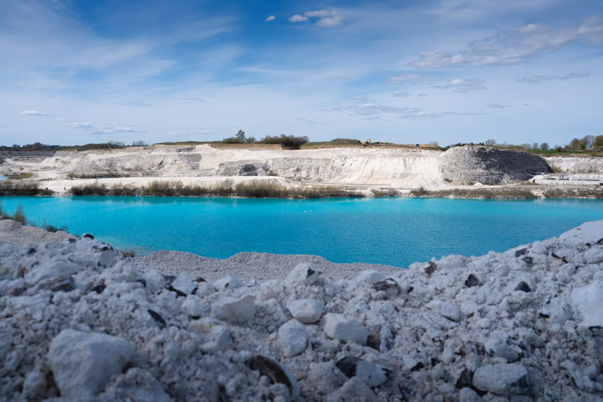 A quarry cut into pale limestone, with bedded rock faces above a flooded pit of bright turquoise water and broken white rock in the foreground