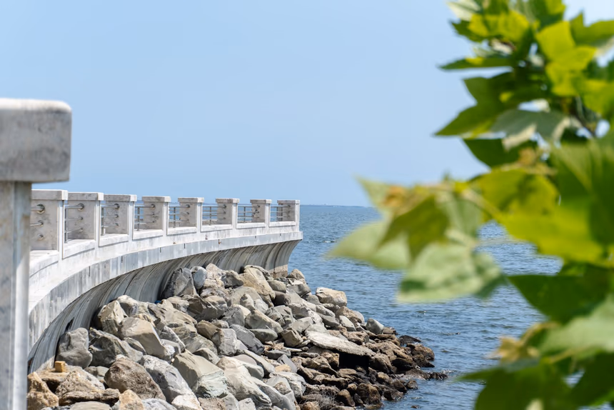 A curving concrete seawall with a capped top and railing, with a bank of large rip-rap boulders piled against its base where it meets the water