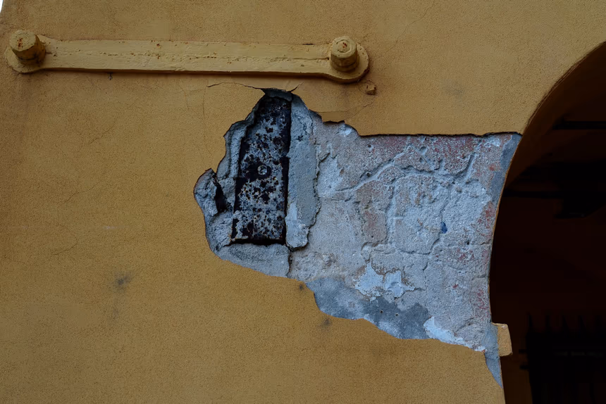 Stucco and concrete broken away from a wall face, revealing a heavily corroded flat steel member embedded in the wall behind