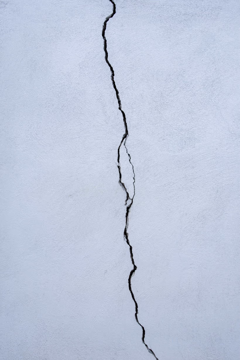 A crack running the full height of a white stucco wall, narrow at the top and wider partway down, with a step where it changes direction
